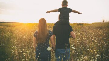 The son is sitting on the father's shoulders and they are looking on the sunset in the field and mother stands near them