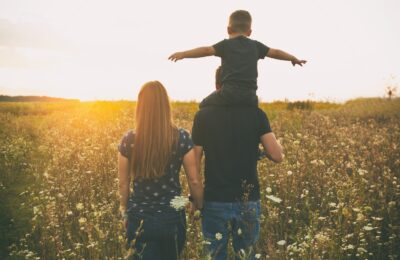 The son is sitting on the father's shoulders and they are looking on the sunset in the field and mother stands near them