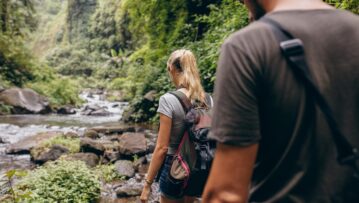 Rear view shot of young man and woman hiking in forest. Tourist couple walking by stream.